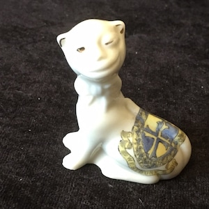 May include: White porcelain figurine of a cat with a winking face and a blue and gold crest on its back. The crest features a cross and the text "ECCLESIA  S.  MARY  THE  VIRGIN  CHUR  1874".