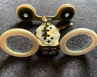 May include: Antique opera glasses with a brass frame and cream-colored accents. The lenses are round, and the frame features decorative cutouts and a central adjustment mechanism. The glasses are designed for viewing performances.