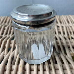 May include: A clear glass jar with a silver lid. The jar has a hexagonal shape and is decorated with vertical lines. The lid has a smooth, shiny surface.