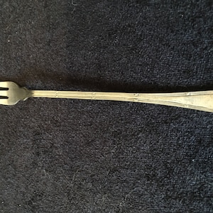 May include: A silver-toned fork with three tines and a long, slender handle. The handle has a decorative pattern.