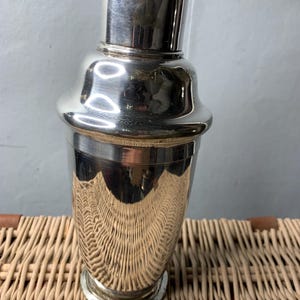 Op de afbeelding: Een gepolijste zilveren cocktailshaker met een afgeronde body en een dop. De shaker reflecteert zijn omgeving. Hij staat op een geweven rieten oppervlak, wat wijst op een klassiek barthema. De hoogte van de shaker is ongeveer 20 cm.