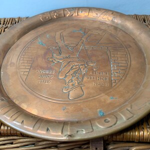 Puede incluir: Una bandeja redonda de cobre con una imagen en relieve de un hombre subiendo una escalera. La bandeja tiene el texto "BORN EIGHTEEN TWENTY STILL GOING STRONG" alrededor de la imagen.