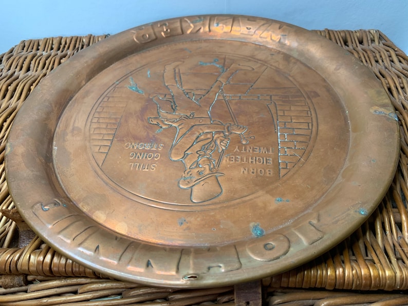 Puede incluir: Una bandeja redonda de cobre con una imagen en relieve de un hombre subiendo una escalera. La bandeja tiene el texto "BORN EIGHTEEN TWENTY STILL GOING STRONG" alrededor de la imagen.