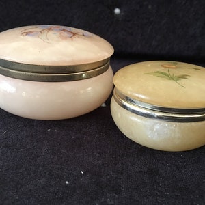 May include: Two round, decorative, alabaster trinket boxes with floral designs. The larger box is pink with a blue and brown floral design. The smaller box is yellow with a green and yellow floral design. Both boxes have a gold metal rim around the lid.