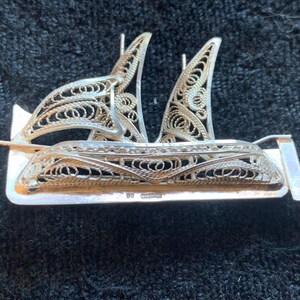 Op de afbeelding: Een zilveren filigrein broche in de vorm van een zeilboot. De broche is versierd met ingewikkelde ontwerpen en heeft een zilveren achterkant.