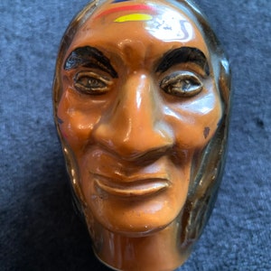 May include: A ceramic figurine of a Native American man's head with a painted face. The face is brown with black eyes, eyebrows, and a mouth. The forehead has a yellow, red, and blue painted design.