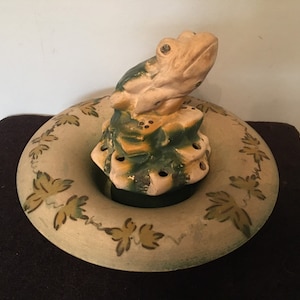 Puede incluir: Una figurita de rana de cerámica se sienta encima de una base de cerámica verde y amarilla con agujeros. La base se encuentra sobre un plato redondo de cerámica más grande con un diseño de hojas verdes y amarillas.