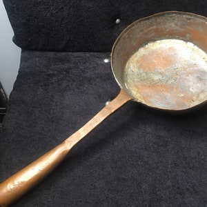 Pode incluir: Uma panela de cobre vintage com uma longa pega de madeira. A panela tem um aspeto desgastado e envelhecido e é perfeita para cozinhar em fogo aberto.