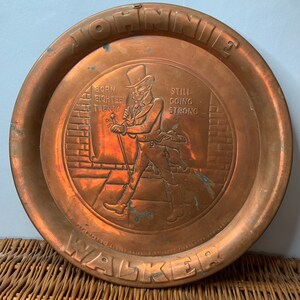 Puede incluir: Una bandeja de servir de cobre con una imagen en relieve de un hombre con un sombrero de copa caminando. La bandeja est&aacute; estampada con las palabras "Johnnie Walker" y "Born 1820 Still Going Strong".