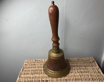 May include: A vintage bell with a wooden handle and a brass bell. The handle is a dark brown color, and the bell has a reddish-brown and gold finish. The bell is sitting on a woven wicker surface.