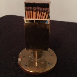 May include: A vintage brass match holder with a round base and a rectangular top. The base is decorated with small coloured dots. The matchbox is open, revealing the matchsticks with brown tips.
