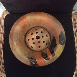 Puede incluir: Un jarrón redondo de cerámica marrón con un diseño floral. El jarrón tiene una base circular elevada con agujeros para sujetar las flores. El diseño presenta hojas negras y naranjas sobre un fondo marrón.