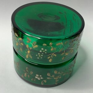 Puede incluir: Un tarro de vidrio verde esmeralda de dos piezas con tapa. El tarro está decorado con patrones florales y de hojas doradas. El tarro es redondo y tiene un acabado brillante.