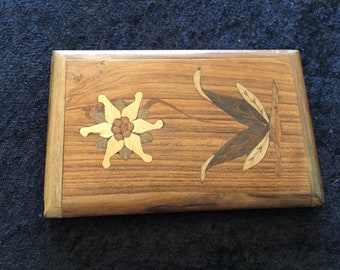May include: A wooden box with a floral design inlaid in the top. The design features a white flower with brown and yellow accents and a brown leaf with yellow accents.