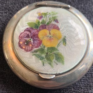 以下が含まれることがあります： 花柄のデザインが施された丸い銀色のコンパクト。蓋には、エンボス加工された白い背景に、紫と黄色のパンジーと緑の葉が描かれています。コンパクトはヒンジで開きます。