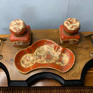 May include: A vintage desk set featuring two red and cream inkwells with floral lids, a matching decorative tray, and a wooden tray with brass handles. The set has a classic, ornate design.