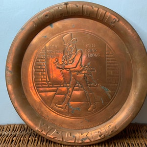 Puede incluir: Una bandeja redonda de cobre con una imagen en relieve de un hombre con un sombrero de copa caminando por una calle. La bandeja tiene las palabras "Johnnie Walker" y "Born 1820 Still Going Strong" alrededor del borde.