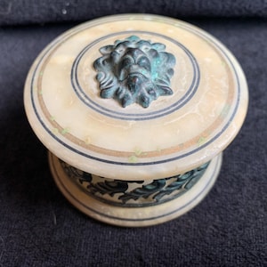 Puede incluir: Una caja de baratijas redonda y antigua con un acabado de mármol color crema. La tapa presenta una cabeza de león de bronce y adornos decorativos. La base tiene un diseño floral de bronce a juego. Un recipiente de almacenamiento decorativo.