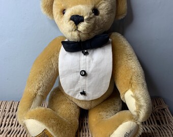 May include: A tan teddy bear wearing a white tuxedo with a black bow tie. The bear is sitting on a wicker chair. The bear's paw has a label that reads "Steiff" and "Made in Germany". The bear's other paw is signed "Paul & Emma"