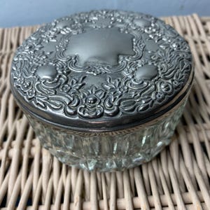Puede incluir: Un tarro de vidrio transparente con una tapa de plata con un diseño floral ornamentado. La tapa tiene un centro elevado con una superficie lisa y reflectante.