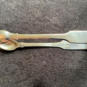 Könnte beinhalten: Silberne Zuckerzange mit einem löffelartigen Ende. Die Zange ist aus poliertem Silber und hat ein schlichtes Design.