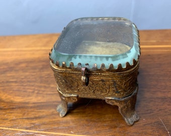 May include: A small, ornate, gold-coloured trinket box with a glass lid and three legs. The box has intricate floral designs.