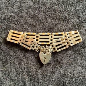 Puede incluir: Collar gargantilla plateado con cierre de candado en forma de corazón. El collar presenta una serie de barras horizontales conectadas por barras verticales. El candado en forma de corazón tiene un ojo de cerradura.