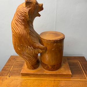 May include: A wooden bear figurine holding a wooden cylinder with a lid. The bear is standing on a wooden base. The figurine is carved in a rustic style.