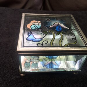 May include: A silver-toned trinket box with a glass top and bottom. The top features a stained glass design of blue, pink, and green flowers.