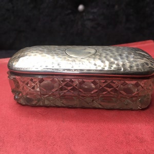 May include: A silver and glass trinket box with a hammered lid and a clear glass base with a diamond pattern. The lid has a circular inscription.