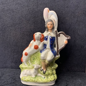 Puede incluir: Una figura de cerámica que representa a un hombre con abrigo azul y calzones blancos, tocando un arpa. También hay un perro con manchas rojas y un cordero. La figura está sobre una base verde con un elemento decorativo blanco y rosa.