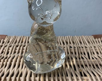May include: A clear glass cat figurine with a seated pose. The cat has visible bubbles within the glass and is sitting on a woven wicker surface. The figurine has a simple, yet charming design.