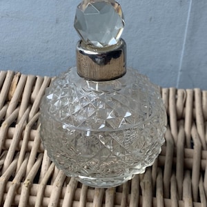 May include: A clear, round perfume bottle with a faceted crystal stopper and a silver-colored metal collar. The bottle has a diamond-cut pattern and sits on a woven wicker surface. A vintage-style decorative item.