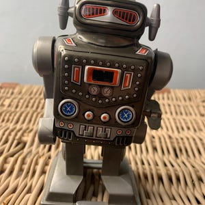 May include: A metallic gray toy robot featuring a rounded head with orange-accented eyes, two side antennas, and a torso decorated with printed dials, gauges, and buttons. The robot stands on two rectangular feet and has a wind-up key on its side.
