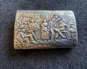 Op de afbeelding: Zilveren rechthoekige doos met een gedetailleerde reliëfscène van mensen in een bijeenkomst. De figuren zijn gekleed in kleding uit die tijd, sommigen zitten aan tafels en anderen staan. De doos heeft een licht aangeslagen uiterlijk.