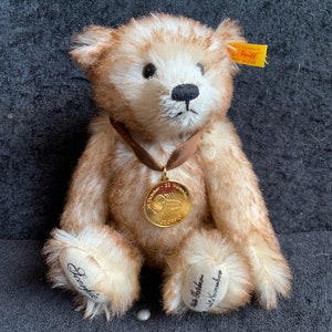 May include: A plush teddy bear with light brown fur and a black nose. The bear wears a brown ribbon with a gold medallion that reads "14 October - 22 November Scorpio". The bear's paws are embroidered with the name "Sophie" and the dates.