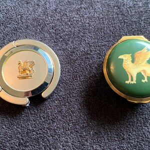 May include: Two round, decorative compacts. One is silver with a gold griffin design. The other is green with a gold griffin design.