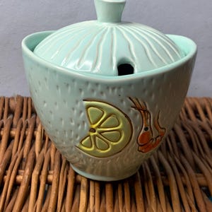 May include: A light blue ceramic sugar bowl with a lid. The bowl features a lemon and a shrimp design. The lid has a small hole in the top.