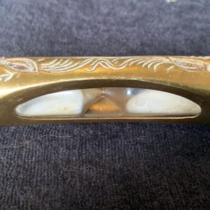 May include: A vintage hourglass with a brass casing and clear glass windows. The brass is etched with floral designs. The hourglass contains white sand. The item is resting on a dark, textured surface.