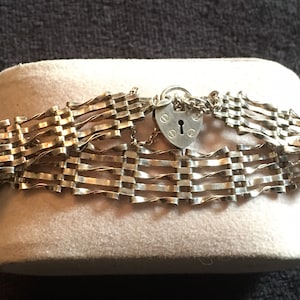 Könnte beinhalten: Ein silbernes Kettenarmband mit einem herzförmigen Schlossverschluss. Das Armband besteht aus einem gewebten Kettenmuster.