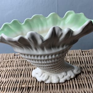 May include: A white ceramic seashell-shaped vase with a pale green interior. The vase has a textured, spiralled base and a ruffled rim. The vase is displayed on a woven wicker surface.