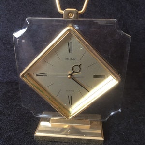 May include: A gold-toned Seiko quartz clock with a diamond-shaped face and a clear acrylic case. The clock has a handle on top and sits on a gold-toned base.
