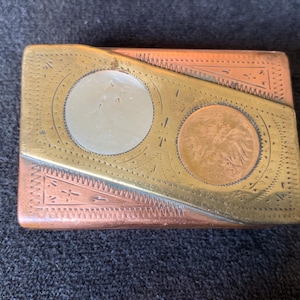 Puede incluir: Una caja metálica rectangular con acabado en cobre y latón. La caja presenta dos incrustaciones circulares, una plateada y otra cobriza, y está decorada con patrones grabados. La caja es probablemente vintage o antigua.