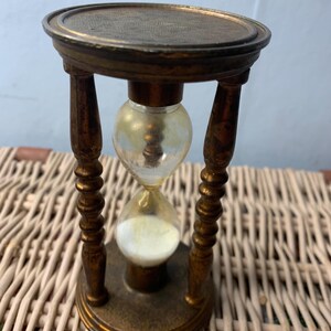 May include: An antique hourglass with a brass frame and a clear glass bulb. The hourglass has a decorative top and base, and the wooden supports are intricately carved. The sand inside is white. A classic timekeeping device.