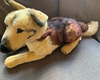 May include: A plush dog toy featuring a tan face with dark brown accents around the ears and muzzle, and a dark brown and black patterned back. The stuffed animal is in a lying position with soft fur, dark eyes, and a black nose, crafted with realistic coloring.