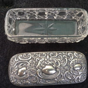 Puede incluir: Una caja rectangular de plata y vidrio con un diseño ornamentado. La parte superior de la caja está hecha de vidrio transparente con un patrón de vidrio cortado. La parte inferior de la caja está hecha de plata con un intrincado diseño floral.
