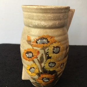 May include: A beige ceramic vase with a floral design. The vase has a rounded body and a flared top. The flowers are painted in shades of yellow, orange, and brown.