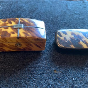 May include: Two small, ornate boxes made of tortoiseshell. The larger box has a hinged lid and a silver latch. The smaller box is rectangular and has a flat lid.
