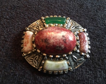 May include: A silver Celtic knot brooch with a red and white stone in the center and four smaller stones around it. The smaller stones are white, red, and green.