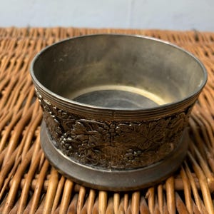 May include: A silver-toned metal wine coaster with a detailed embossed grape and leaf design around the base. The coaster has a circular shape with a slightly raised rim and sits on a woven wicker surface.